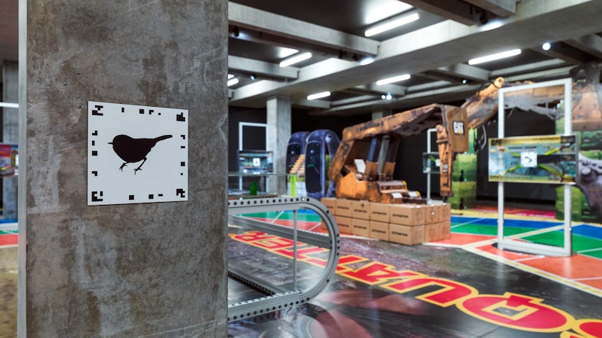 Gallery with large board game-style floor mat pieces shaped like mining machinery. Foreground shows QR code on concrete pillar.