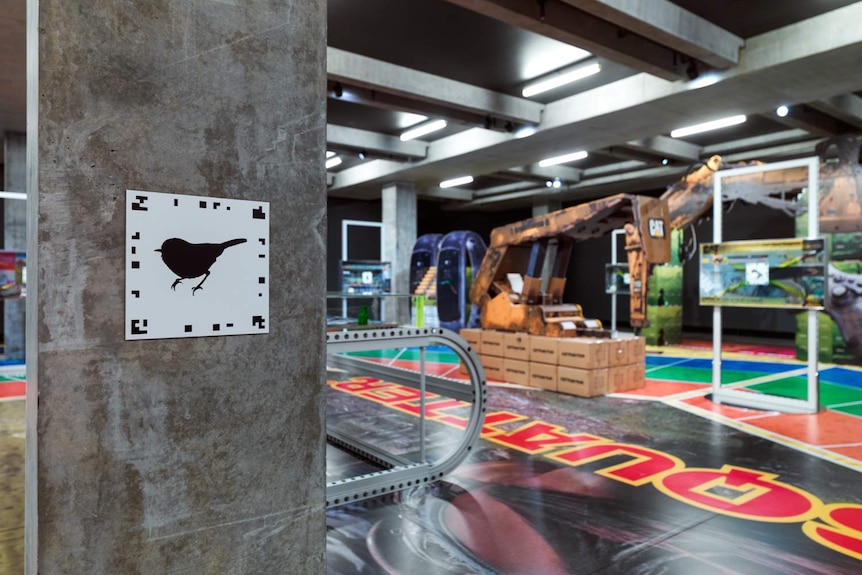 Gallery with large board game-style floor mat pieces shaped like mining machinery. Foreground shows QR code on concrete pillar.