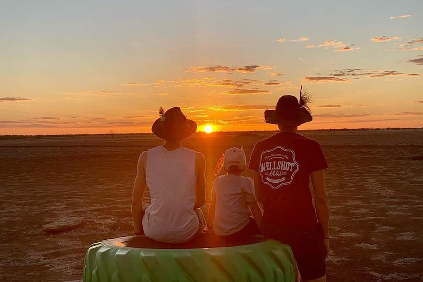Three young people watch the outback sunset while sitting on a tractor tyre  
