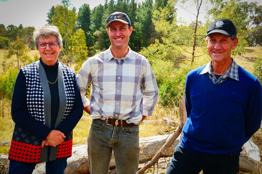 A man in the middle with a older woman on his right and an older man on his left with a vegetation in the bcakground.