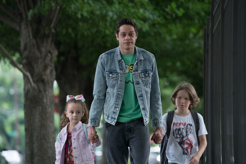 A young man wearing denim jacket looks unimpressed as he walks down leafy sidewalk holding the hands of a female and male child.