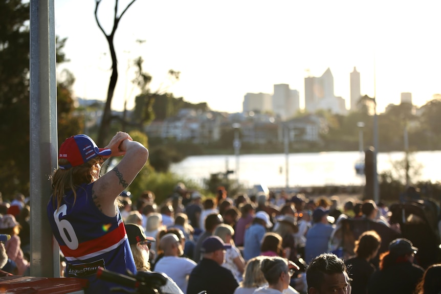 A man with a mullet and arm tattoo in a Bulldogs guernsey leans against a pole and 100s of people sit before the river and city
