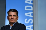 ASADA boss David Sharpe stands, arms folded, in front of a blue sign with ASADA written in white letters