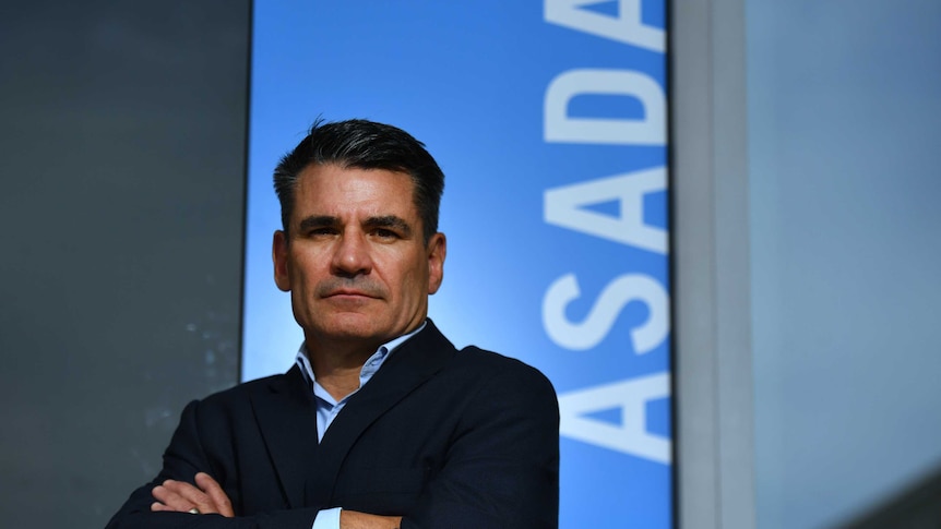 ASADA boss David Sharpe stands, arms folded, in front of a blue sign with ASADA written in white letters