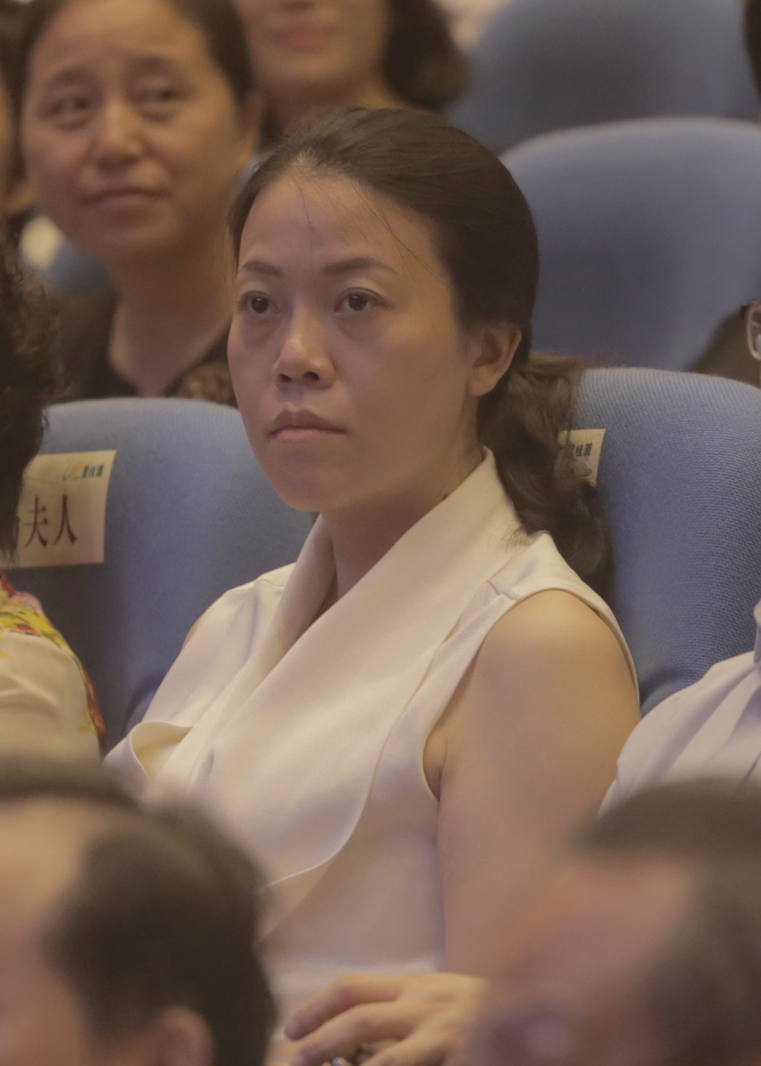 A woman wearing a white collared waistcoat sits in an auditorium crowd