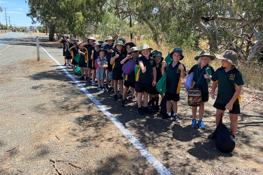 A line of primary school kids standing next to a line on the side of the road. 
