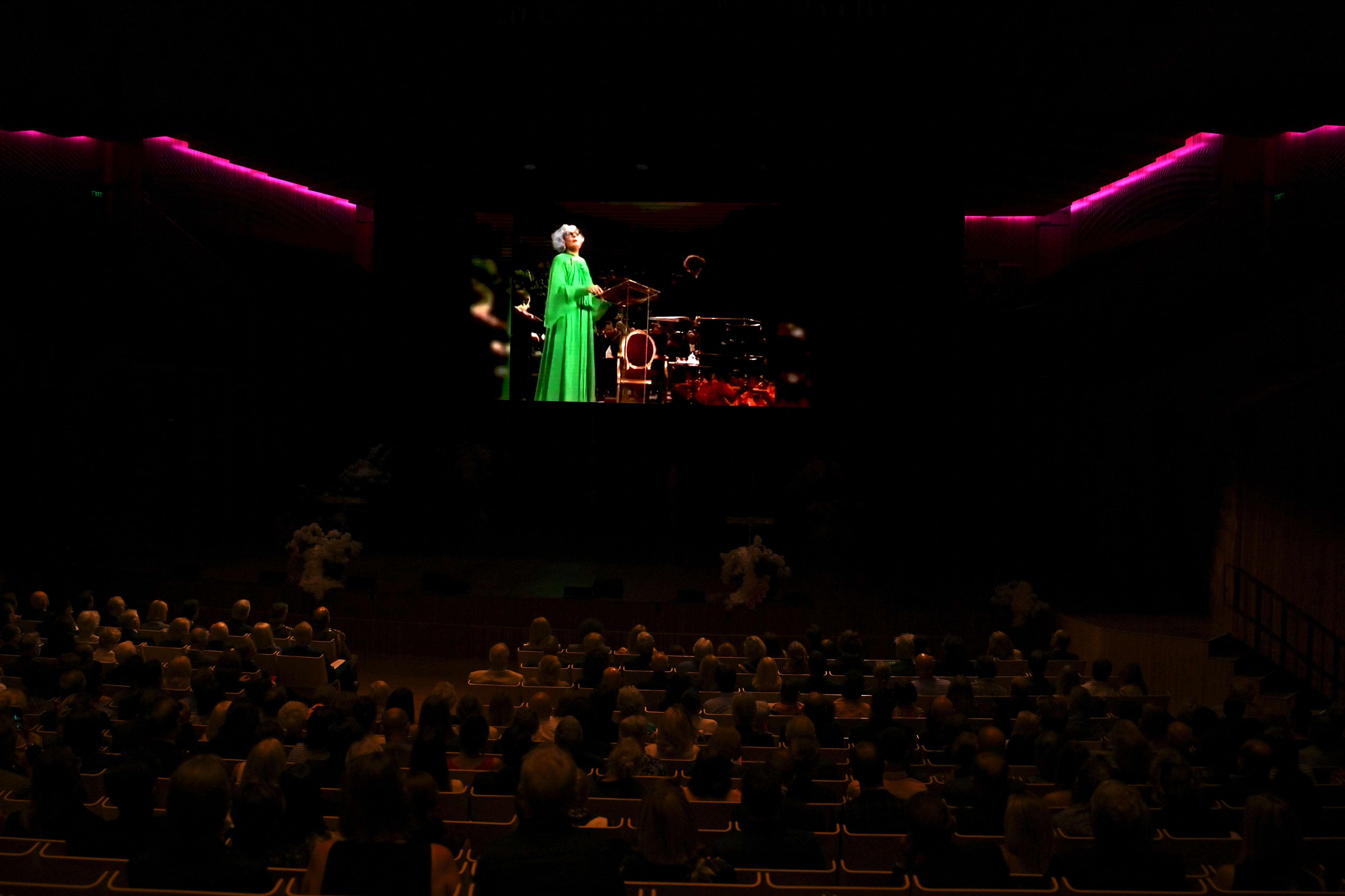 A crowd of people watch a video inside the Opera House of Dame Edna singing.