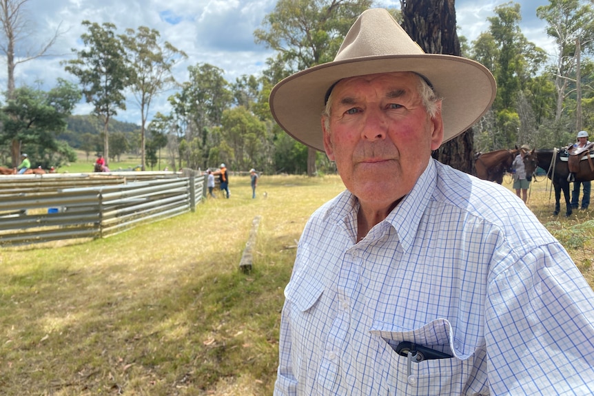 A man in a white checked shirt with a broad-brim hat stands in the foreground, horses and an arena lie behind him. 