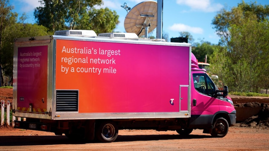 A bright pink Telstra van with the words "Australia's largest regional network" on the side, driving in a remote area.