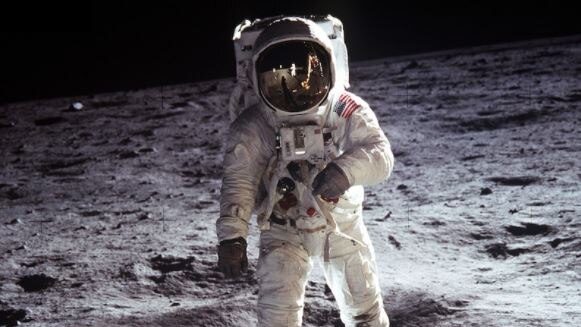 An astronaut stands on the surface of the Moon, the lunar module and another astronaut are visible in his helmet's reflection.