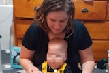 A woman sits on the floor with her baby son, whose face is pixellated.