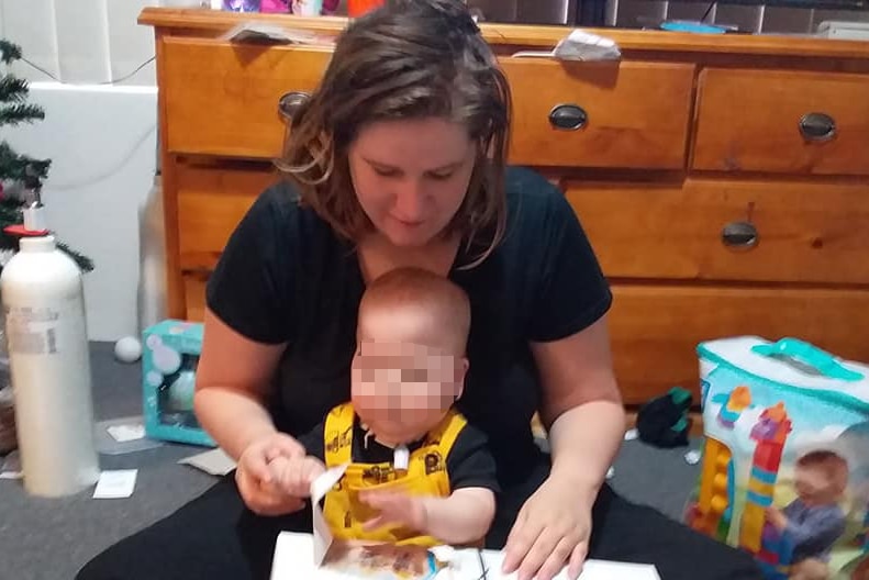A woman sits on the floor with her baby son, whose face is pixellated.