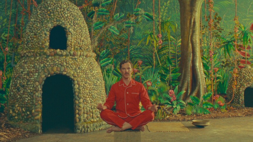 Benedict Cumberbatch in red pyjamas sits in a cross legged meditation position in front of a bright background