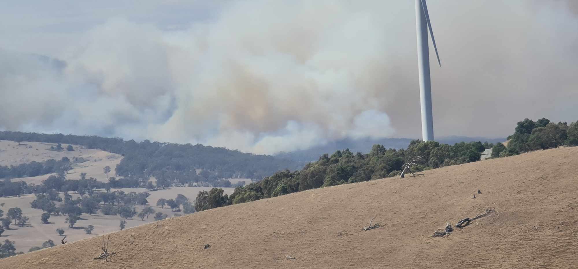 Smoke billowing behind a hill with a wind turbine on it.