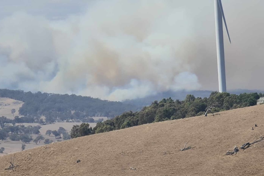 Smoke billowing behind a hill with a wind turbine on it.