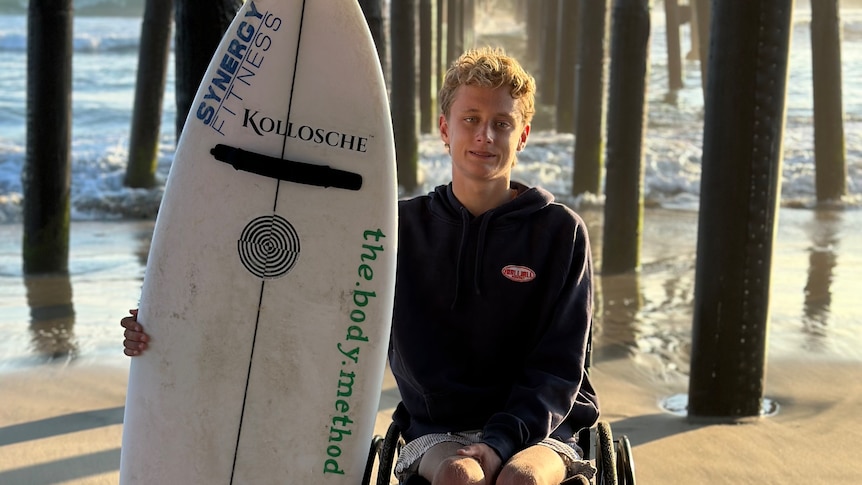 A teenager boy with a surfboard on a beach under an ocean pier