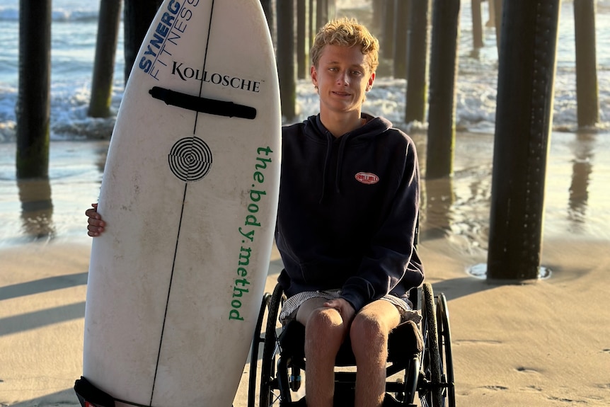 A teenager boy with a surfboard on a beach under an ocean pier