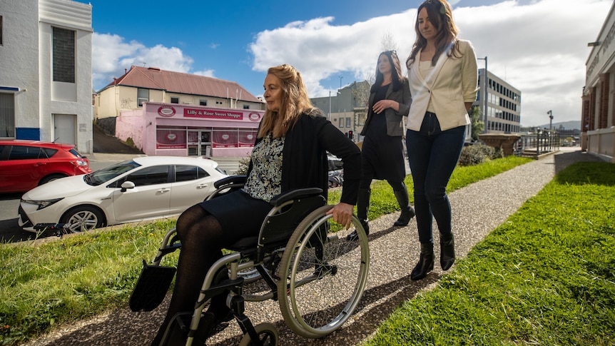 A woman in a wheelchair makes her way down a path as two younger women follow.