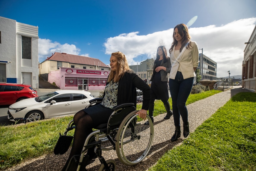 A woman in a wheelchair makes her way down a path as two younger women follow.