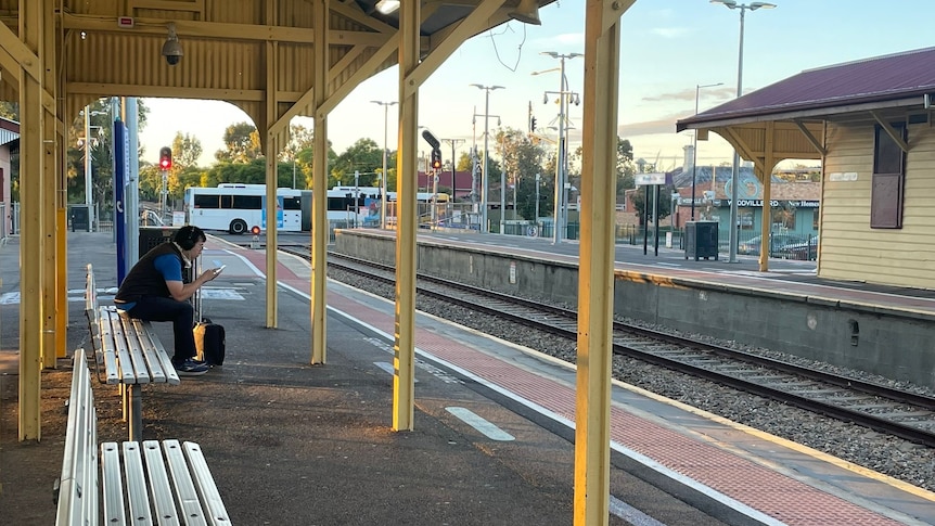 man with headphones on sits looking at his phone at train station awaiting train