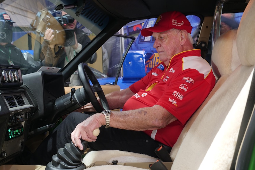 An older man in a cap and branded polo shirt sits behind the wheel of an old racing sedan.