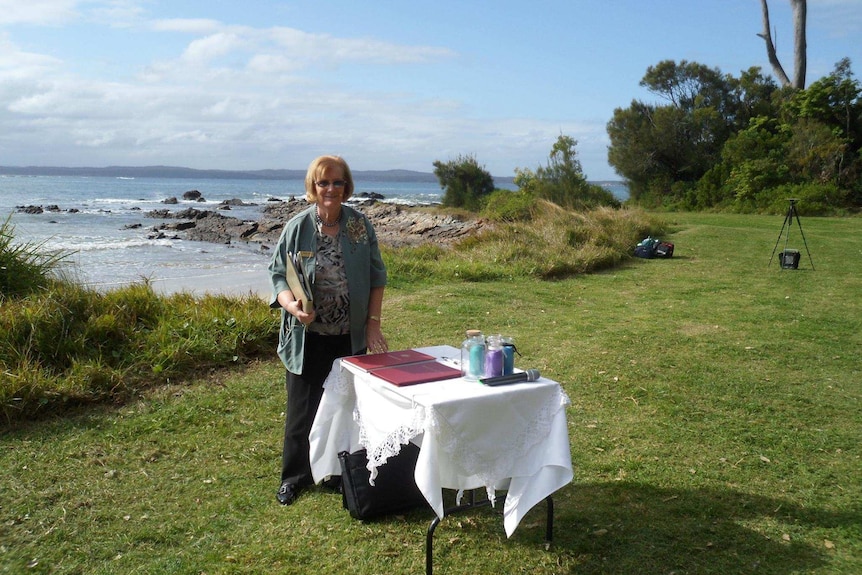 Woman standing behind a table with wedding papers on a grassy area by the sea.