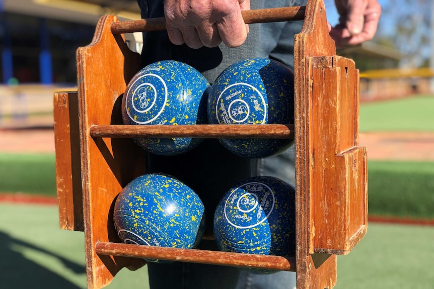 A hand is holding a wooden carry case holding four lawn bowls. They are blue with yellow speckles.