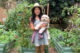 A woman holding a dog stands in front of vegetable beds