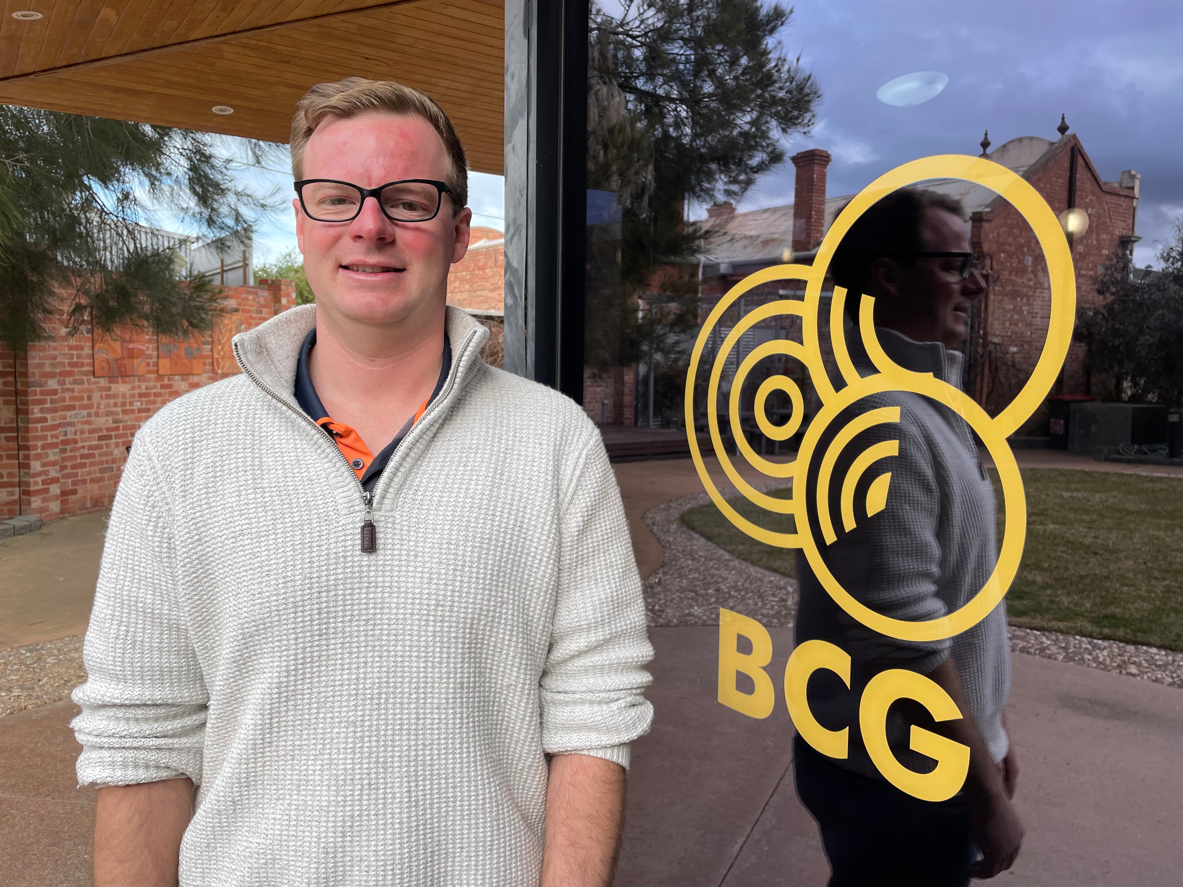 A man with short fair hair, wearing a jumper, stands beside a window with a logo and "BCG" written on it.