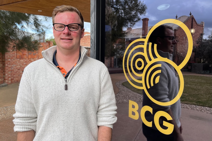A man with short fair hair, wearing a jumper, stands beside a window with a logo and "BCG" written on it.