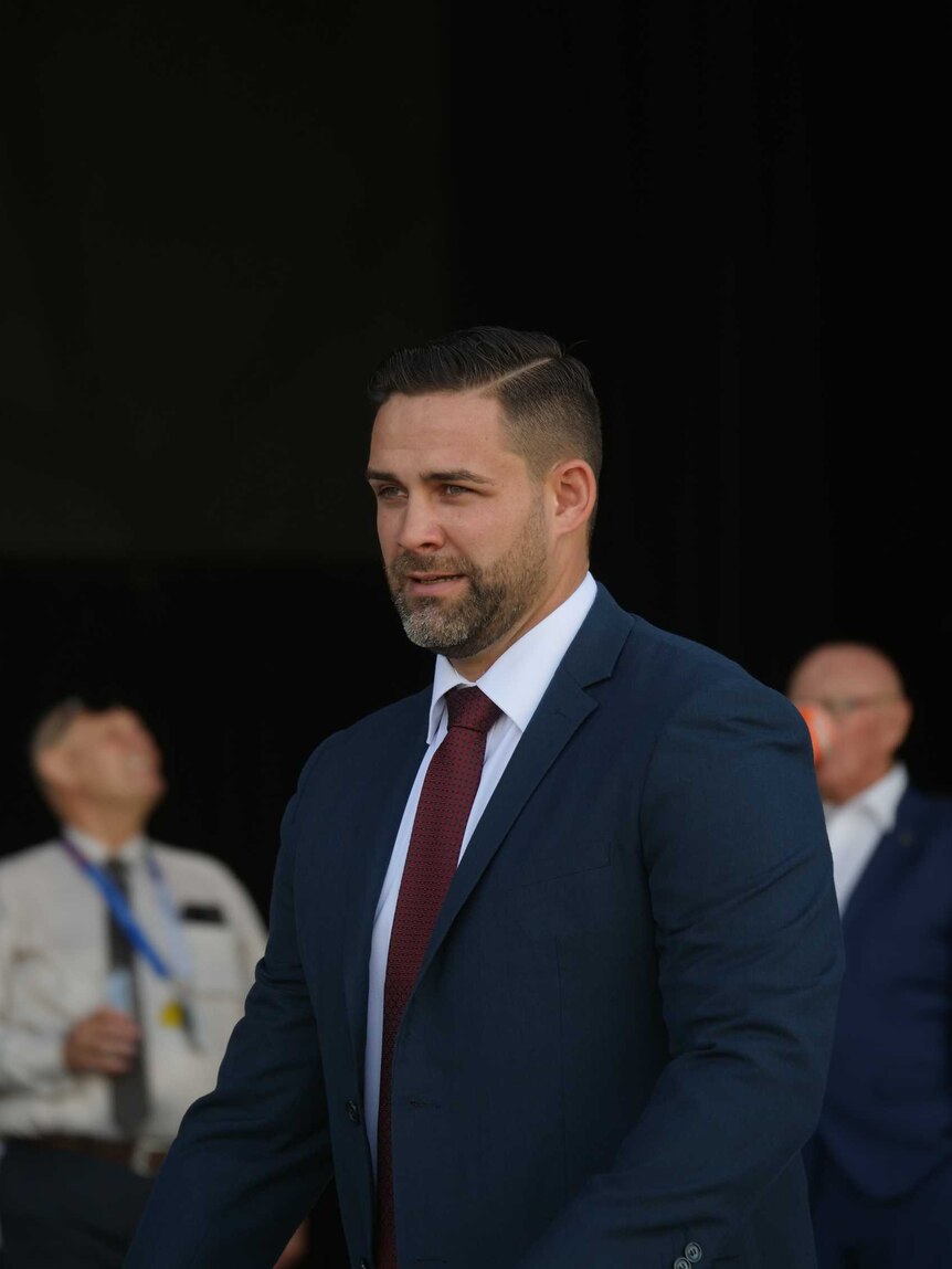 Bearded man wearing a blue suit and maroon tie walking in front of an crowd. 