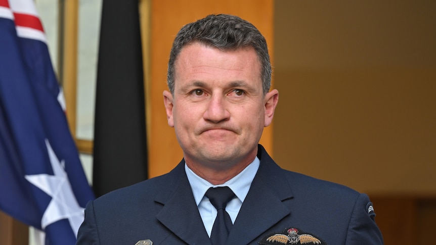 A middle-aged man in a blue air force uniform looks grim in front of the side doors to Parliament House.