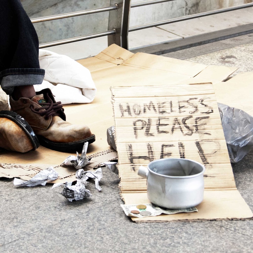 Bottom half of a man sitting on cardboard on the ground wearing worn out boots. In front a sign reads “homeless please help.”