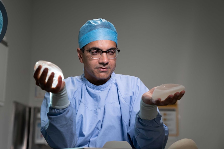 A man rubs me standing in an operating theater holding a breast implant in each hand.