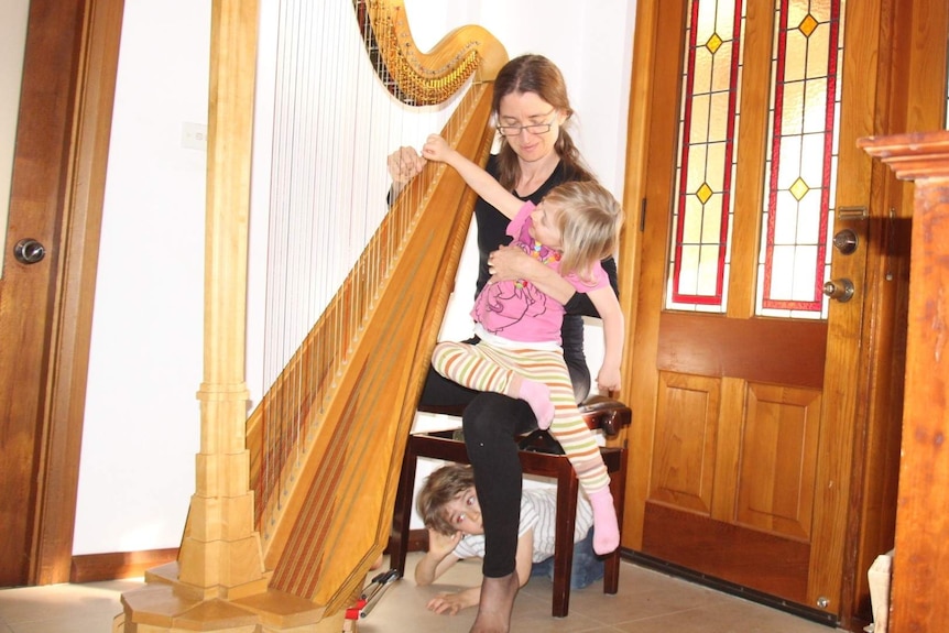 A woman sits playing the harp with a child climbing on her knee and another hiding under her chair.