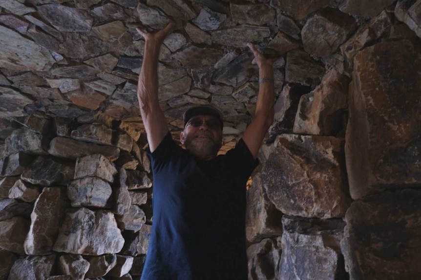 A man in a dark blue shirt and jeans presses his hands to the roof made of rocks in a cave underneath a waterfall.