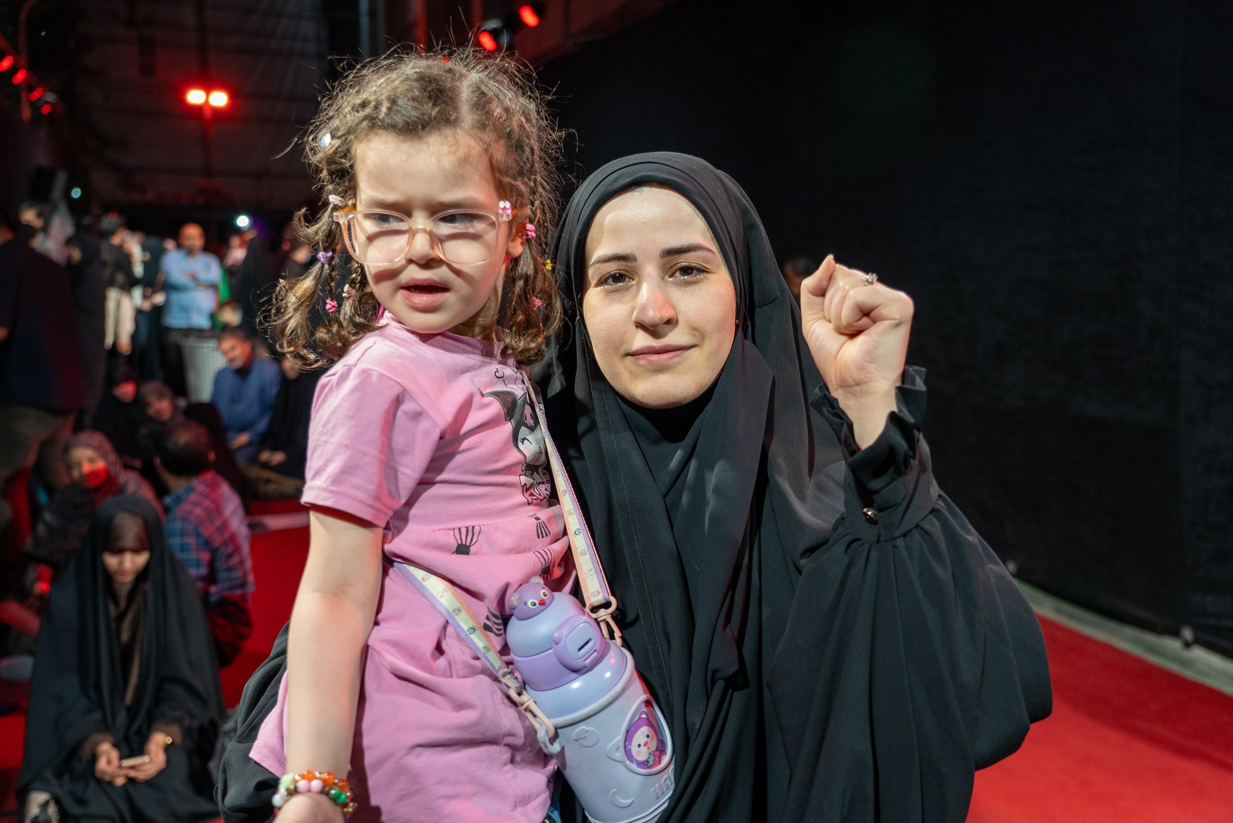 A woman in a black chador holding her daughter. She has her left fist raised.