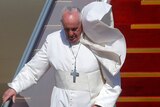 A windswept pope in traditional garb holds railing and walks down steps from plane on sunny day.