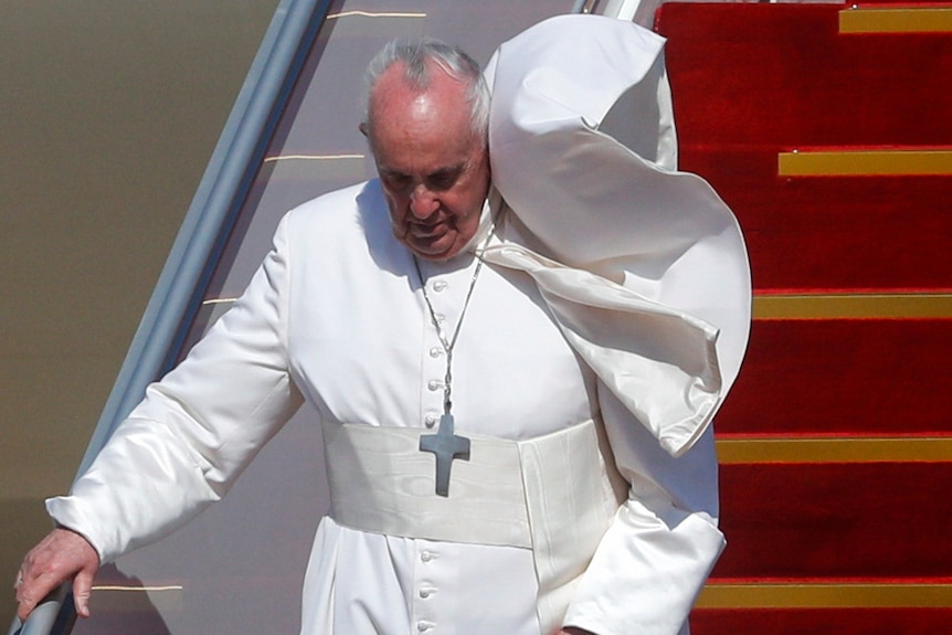 A windswept pope in traditional garb holds railing and walks down steps from plane on sunny day.