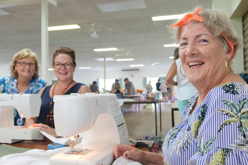 Woman with a bow in her hair and two other women smiling sitting behind sewing machines