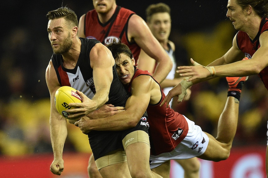 An AFL player holding a football, being tackled from behind by another player.