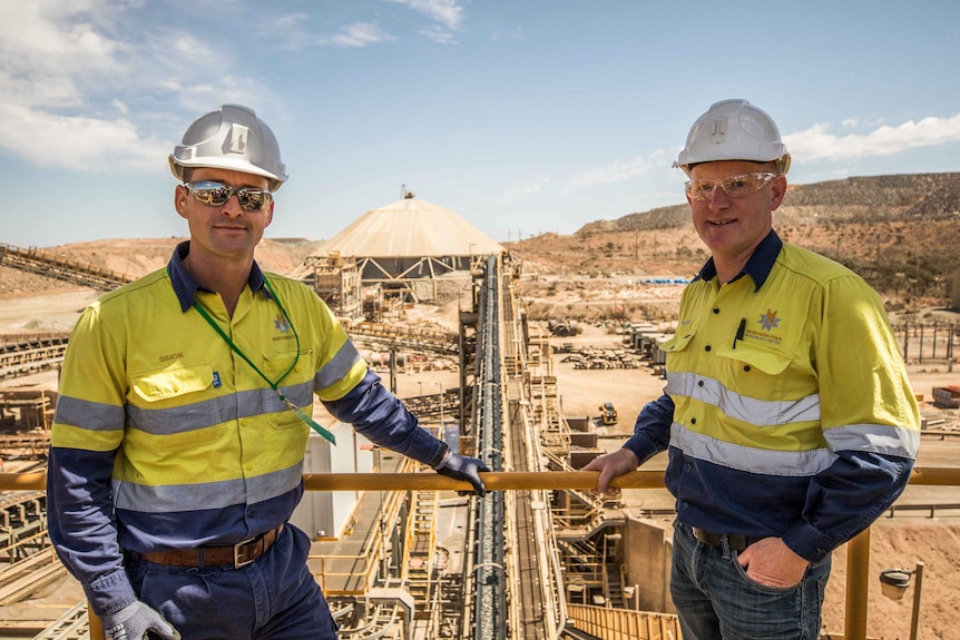 Two men wearing hard hats and high-vis mining workwear on top of a viewing platform of a gold mill.