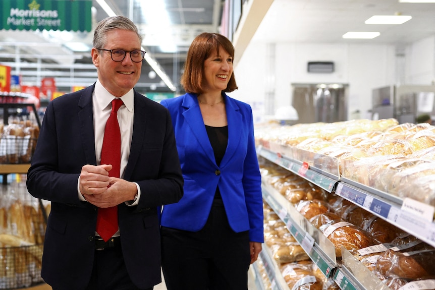 Keir Starmer in suit with red tie and Rachel Reeves in blue blazer walk through a supermarket past shelves of bread