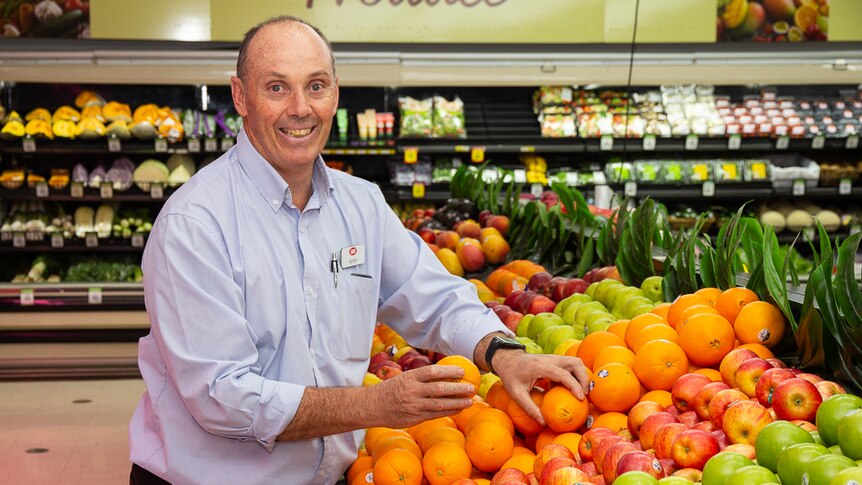 A man in a long sleeve business shirt stands at a fruit stand inside a grocery store, handling oranges