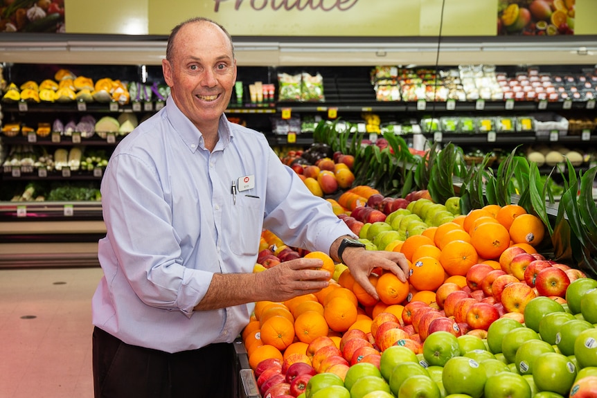 A man in a long sleeve business shirt stands at a fruit stand inside a grocery store, handling oranges