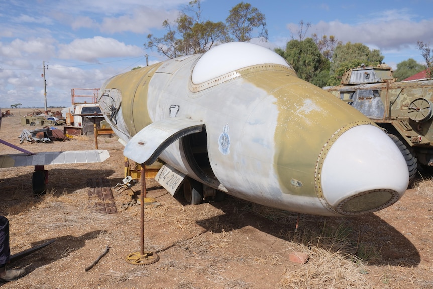 La nariz de un avión de camuflaje gris y verde sin alas se asienta sobre hierba seca con un tanque detrás 