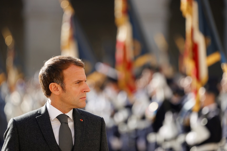 Emmanuel Macron wears a red flower pinned to a suit while looking over his shoulder at armed guards.