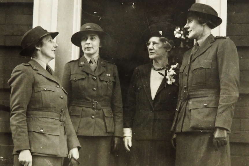 Four women in army uniforms stand outside a building entrance chatting.