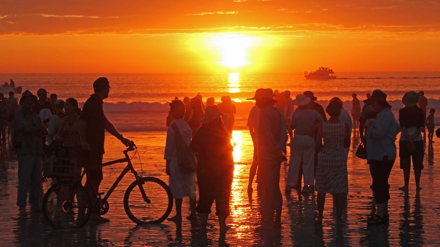 Tourists and bike riders on a beach watching the sun set over the ocean.