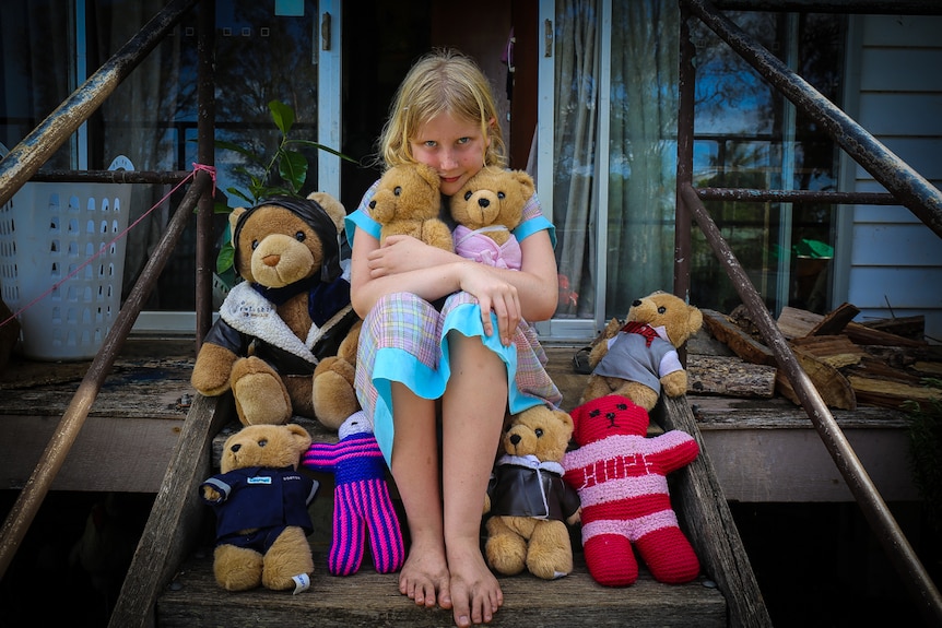A blond girl sitting on steps cuddling teddy bears.